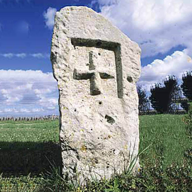 A weathered stone marker with a carved cross standing in a green vineyard under a blue sky with white clouds, Pomerol, Bordeaux.